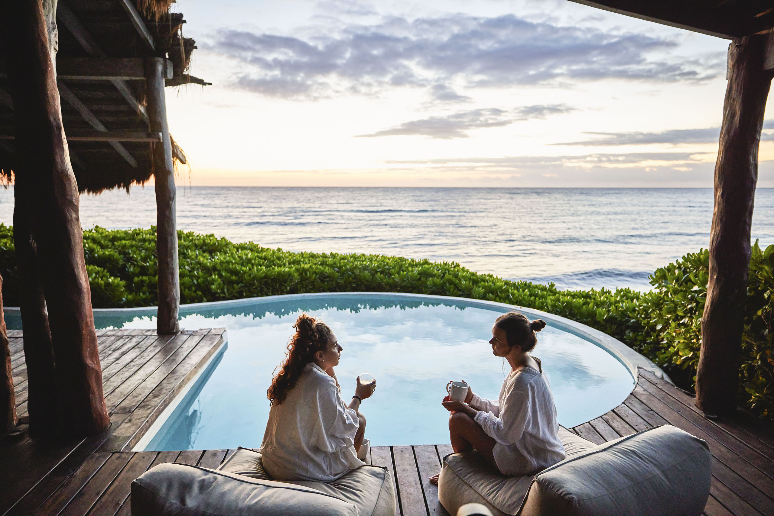 Wide shot of female friends in discussion while sitting poolside at luxury suite of tropical resort at sunrise